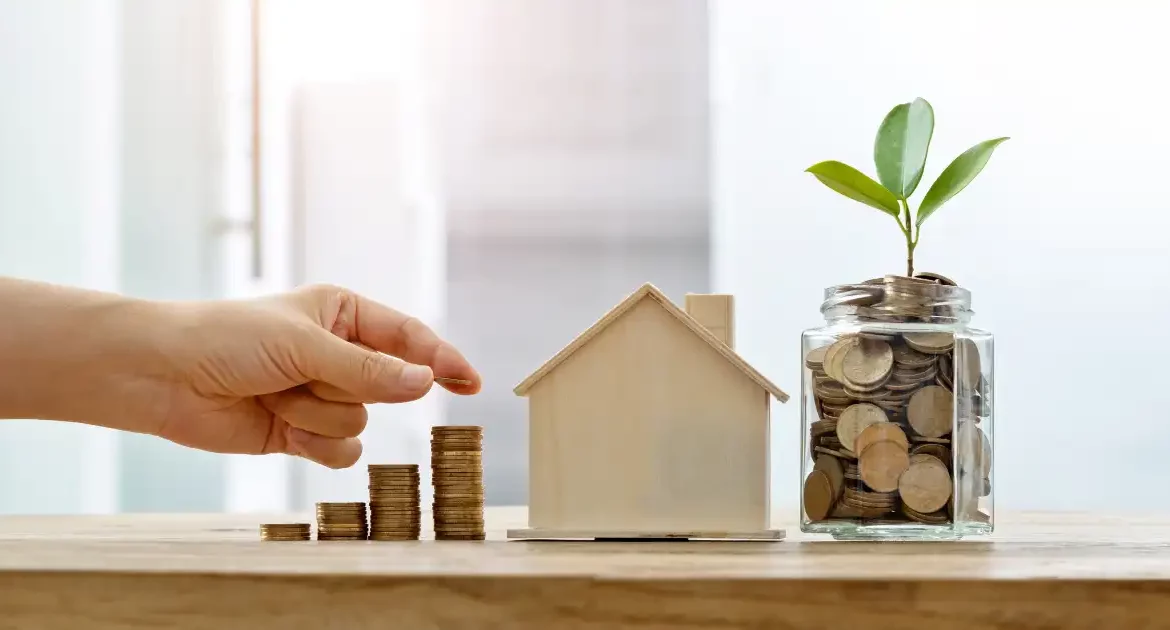 Hand placing stacks of coins in ascending order next to a wooden house and a plant in a coin jar.